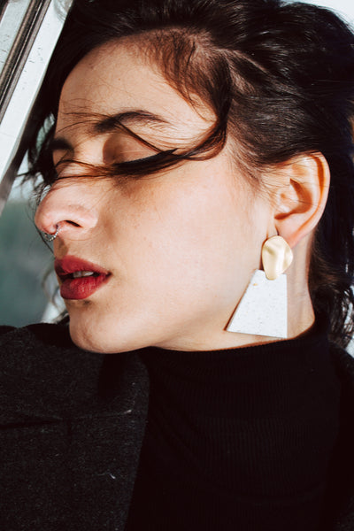 Close up of model'd face wearing black clothes with statement white and gold trapezoid earrings